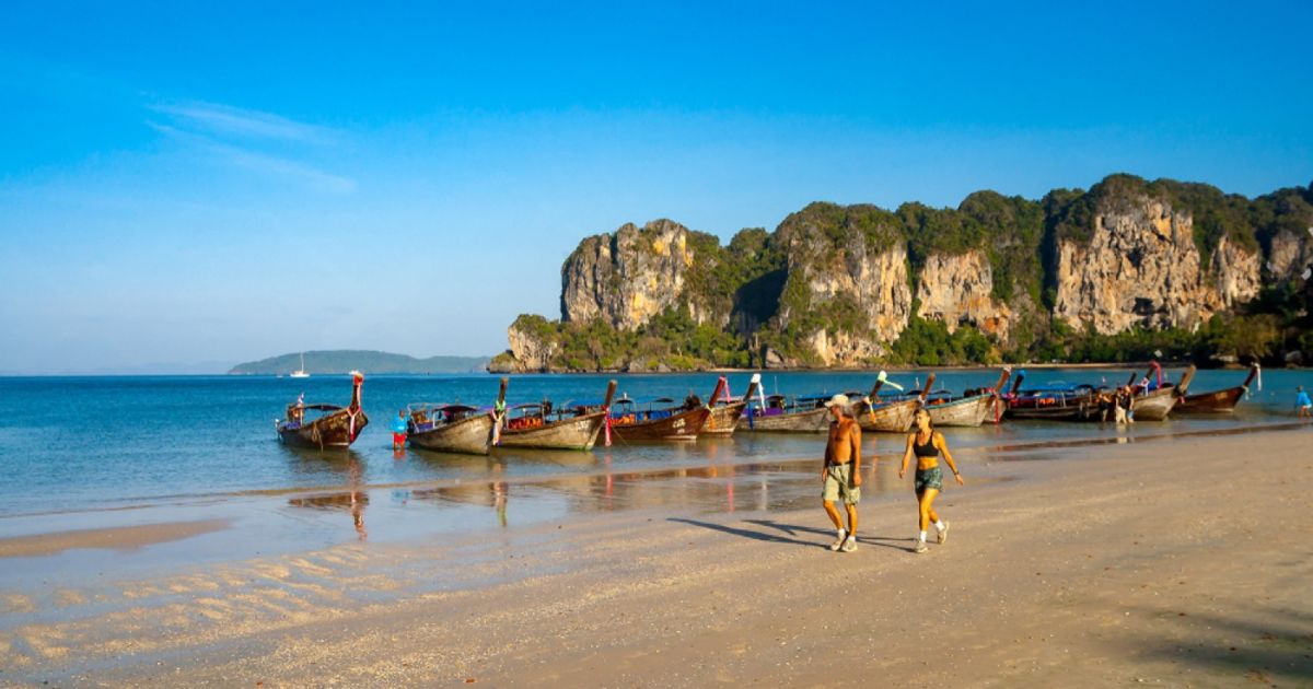 Two people walking along Railay beach in Krabi at sunrise, longtail boats moored beside the limestone cliffs